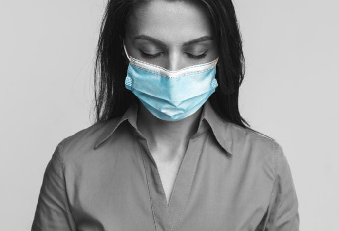 Mujer triste, deprimida, en blanco y negro con cubre bocas azul cerrando los ojos y llorando durante la pandemia de coronavirus